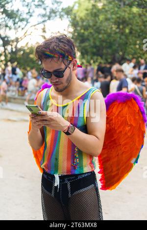 Junger Mann mit leuchtender Sonnenbrille in mehrfarbigen Kleidern und Accessoires, der sein Handy benutzt, während er während des LGBTQ+ Stolz im Park steht Stockfoto