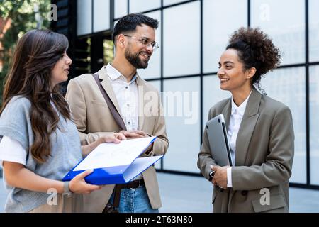 Vielfältige Gruppe junger Geschäftsleute außerhalb des Bürogebäudes, diskutieren und sprechen Männer und Frauen, die den neuen Bauplan lösen, Team von Unternehmen e Stockfoto