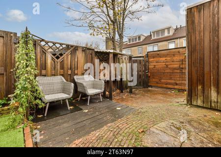 Korbstühle, die durch einen Holzzaun in einer gepflasterten Terrasse vor bewölktem Himmel im Hinterhof des modernen Hauses platziert werden Stockfoto