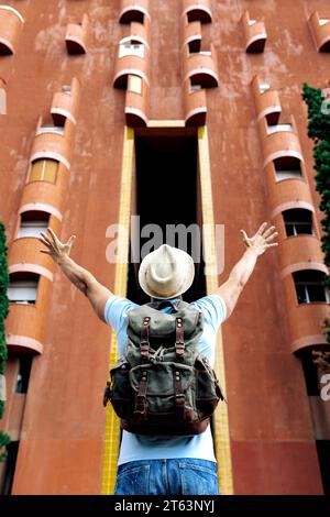 Flacher Blick auf einen Touristen von hinten mit Hut und Rucksack, der mit ausgestreckten Armen vor einem unverwechselbaren hohen Gebäude in Barcelona steht Stockfoto