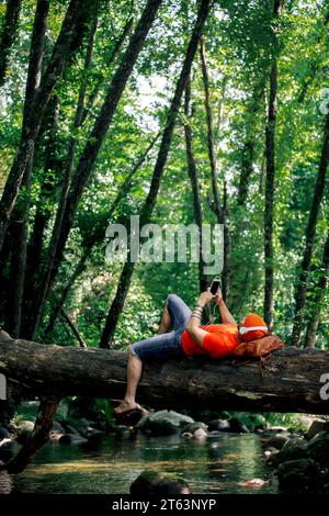 Wanderer lehnt sich auf einem Baumstamm über einem Bach, vertieft in Musik mit seinem Smartphone. Stockfoto