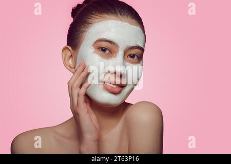 Nahporträt eines jungen asiatischen Modells, das eine Gesichtsmaske aus Ton auf einem rosa isoliert aufträgt. Stockfoto