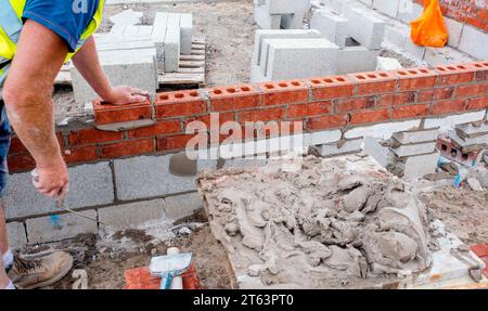 Hart arbeitender Maurer legt Ziegel auf Zementmischung auf der Baustelle. Bekämpfung der Wohnungskrise durch den Bau von erschwinglicheren Häusern Stockfoto