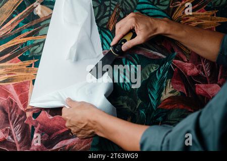 Nahaufnahme eines kaukasischen Mannes in einem grauen Arbeitsmantel mit einem Farbschaber, um die florale Tapete von der Wand zu entfernen Stockfoto