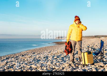 Frau in gelbem Mantel und orangefarbenem Schal geht und entspannt mit gelbem Koffer am Meer bei Sonnenaufgang. Britischer kalter Winter. Lokales Tourismuskonzept. Stockfoto