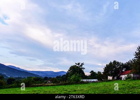 Bauernhäuser und eine grüne Wiese am Hang. Die Sonne hinter den Wolken erleuchtet sowohl den Himmel als auch die Wolken mit romantischem Licht. Die Bergkette Stockfoto