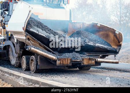 Asphaltfertiger mit heißem Asphalt, der neue Straßenoberflächen auf neuen Wohnbaustellen verlegt, und Straßenarbeiter in orangefarbenem Hi-viz als nächstes Stockfoto