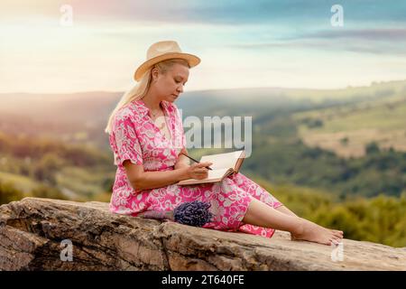 Frau in einem Kleid und einem Hut mit einem Lavendelstrauß in den Händen auf einem Hügel bei Sonnenuntergang im Sommer Stockfoto