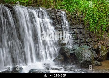 Kleiner Wasserfall mit einem Stück Steinmauer in dichter Vegetation und nassen Steinen Stockfoto