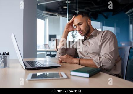 Ein verärgerter, müder junger afroamerikanischer Mann sitzt am Schreibtisch mit Laptop im Büro, müde von der Arbeit, die Hand auf dem Kopf hält, leidet an Kopfschmerzen, gelangweilt im Büro. Stockfoto