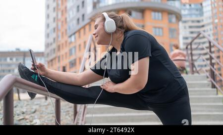 Fette junge Frau, die im Freien Dehnungsübungen macht und Musik mit Kopfhörern hört Stockfoto