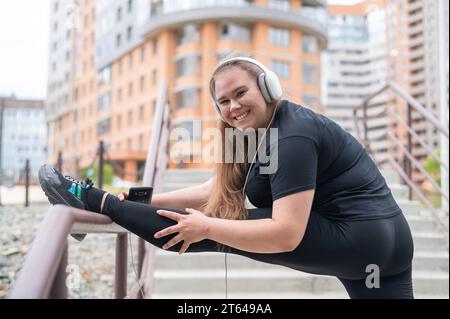 Fette junge Frau, die im Freien Dehnungsübungen macht und Musik mit Kopfhörern hört Stockfoto