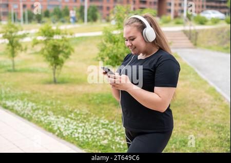 Ein hübsches, fettes Mädchen in schwarzer Sportswear hört Musik auf Kopfhörern und SMS auf dem Telefon, während es draußen läuft. Eine pralle junge Frau ist es Stockfoto