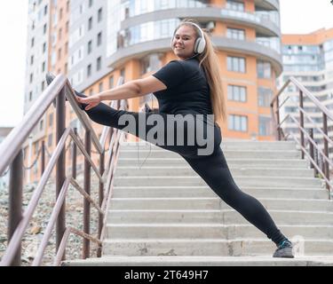 Fette junge Frau, die im Freien Dehnungsübungen macht und Musik mit Kopfhörern hört Stockfoto