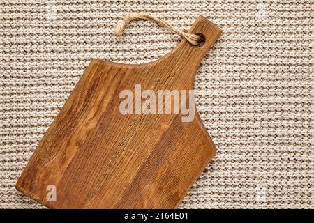 Handgefertigtes braunes Schneidebrett aus Holz auf einer beigen gestrickten Tischdecke. Vorlage für die Präsentation von Speisen auf der Speisekarte. Draufsicht mit Kopierbereich Stockfoto