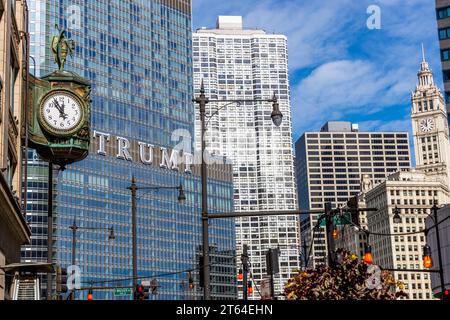 Die Uhr im Juweliergebäude wird kurz vor Hochmittags angezeigt. Im Hintergrund sehen Sie die Schrift Trump aus dem Trump Tower. Chicago, Usa. Trump Tower Chicago auf der DuSable Bridge und gegenüber einer alten Straßenuhr mit der Zeitanzeige „fünf bis zwölf“ Stockfoto