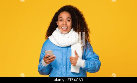 Glückliche schwarze Dame in Winterjacke und Schal mit Telefon gibt die Daumen hoch Stockfoto