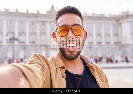 lateinischer Mann mit Bart und Sonnenbrille, der ein Selfie mit seinem Handy macht, mit dem königlichen Palast von Madrid Spanien im Hintergrund Stockfoto