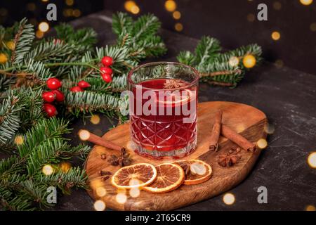 Glühwein in einem Glas auf hölzernem Schneidebrett mit Gewürzen, Fichtenzweigen und Bokeh-Lichtern, Nahaufnahme. Stockfoto