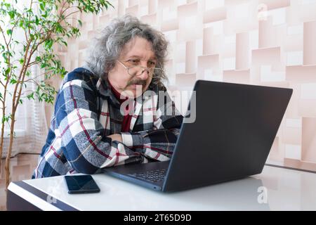 Ein älterer Mann sitzt an einem Tisch und schaut sich ein Video auf einem Computer an. Senior-Mann, der zu Hause am Laptop arbeitet Stockfoto