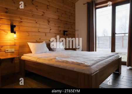Schlafzimmereinrichtung in einem hölzernen Landhaus. Rustikale Blockhütte Schlafzimmer innen, gemütliches Bett durch das große Fenster Stockfoto