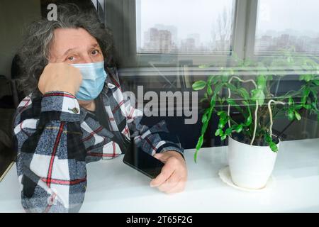 Ein Mann mit Schutzmaske und Telefon in der Hand sitzt am Fenster auf Selbstisolation zu Hause Stockfoto