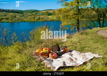 Picknick in der Natur auf einem Hügel mit Blick auf den Fluss. Ein Korb mit Kürbissen, Eicheln, Bananen und Äpfeln liegt auf einem Karo in einer Zelle, daneben befindet sich eine Baske Stockfoto