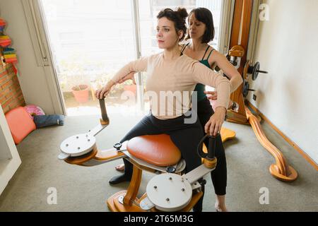 Kaukasische Erwachsene weibliche gyrotonic-Lehrerin unterrichtet weibliche Schülerin zu Hause, hilft ihr bei Körperhaltungen und sitzt auf Maschine, Kopierraum Stockfoto