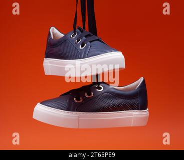 Trendige, legere Schuhe in Marineblau, die vor einem leuchtend orangefarbenen Hintergrund hängen. Perforiertes Leder, dicke Schnürsenkel, Metallösen. Fashion Blog oder Stockfoto