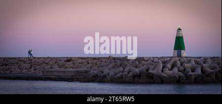 Benalmádena, Spanien - 26. November 2022: Paare, die während des Sonnenuntergangs in Benalmádena zu einem Leuchtturm auf Wellenbrechern gehen Stockfoto