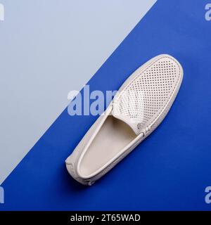 Blick von oben auf den stylischen Mokassinschuh aus perforiertem Leder in Elfenbein, isoliert auf einem kreativen zweifarbigen blauen Hintergrund mit Kopierraum. Kreative Minimalisti Stockfoto