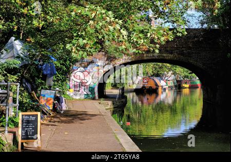 Eine gewölbte Backsteinbrücke über den Regents Canal an der Regents Park Road mit farbenfrohen Graffiti, Kanalbooten und Reflexionen in Camden Town, London, England Stockfoto