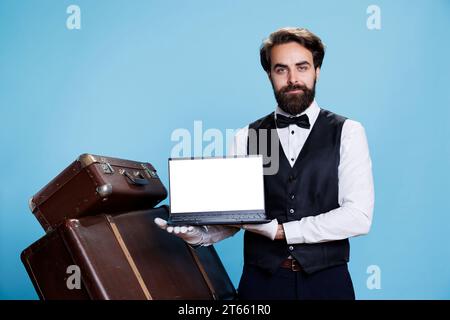 Der Hotelmitarbeiter zeigt einen weißen Bildschirm auf der Kamera und hält einen Laptop mit leerem Copyspace-Display im Studio. Professioneller Luxus-Bellhop mit Krawatte präsentiert Chromakey-Vorlage auf pc, Portier. Stockfoto