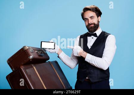 Der Pagen präsentiert einen leeren Bildschirm für die Tourismusbranche vor blauem Hintergrund. Der Concierge des Hotels in formeller Kleidung und Handschuhen hält das Telefon mit weißem Copyspace-Display und posiert vor der Kamera. Stockfoto