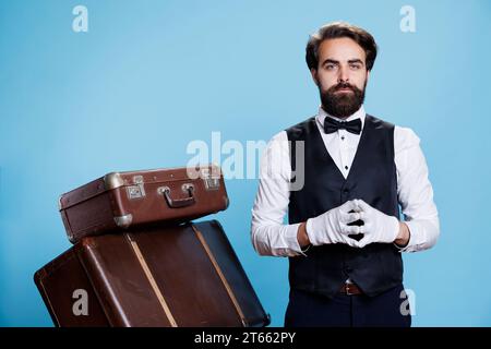 Klassischer Pagen mit Handschuhen und Krawatten, der vor der Kamera posiert, bereitet sich darauf vor, Koffer zu tragen und Hotelgästen Unterstützung zu geben. Junger Mann, der als Türhüter oder Portier arbeitet, Gastgewerbe. Stockfoto