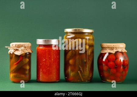 Gemüse in Dosen in geschlossenen Glasgläsern auf grünem Hintergrund. Eingelegte Gurken, Tomaten, Chili-Sauce, Adjika. Scharfer Snack, Hausmannskost. Stockfoto