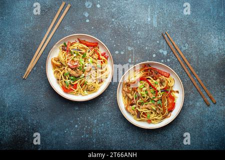 Zwei Schüsseln mit Chow mein oder Lo mein, traditionellen chinesischen Pfannennudeln mit Fleisch und Gemüse, serviert mit Essstäbchen von oben auf rustikalem Blau Stockfoto