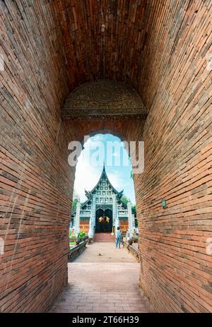 Hoher, schmaler, bogenförmiger, kurzer Tunnel, mit rotem Ziegelwerk gesäumt, führt zu einem verzierten, aus Teakholz gebauten Tempel, mit silberner dekorativer Fassade und Stockfoto