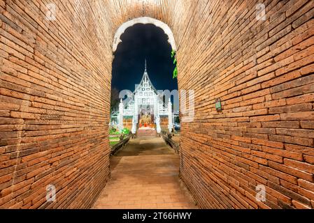 Hoher, schmaler, bogenförmiger, kurzer Tunnel, mit rotem Ziegelwerk gesäumt, führt zu einem verzierten, aus Teakholz gebauten Tempel, mit silberner dekorativer Fassade und Stockfoto