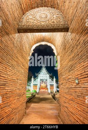 Hoher, schmaler, bogenförmiger, kurzer Tunnel, mit rotem Ziegelwerk gesäumt, führt zu einem verzierten, aus Teakholz gebauten Tempel, mit silberner dekorativer Fassade und Stockfoto