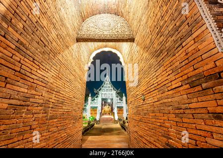 Hoher, schmaler, bogenförmiger, kurzer Tunnel, mit rotem Ziegelwerk gesäumt, führt zu einem verzierten, aus Teakholz gebauten Tempel, mit silberner dekorativer Fassade und Stockfoto