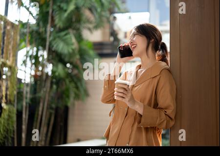 Seitenansicht einer fröhlichen und schönen jungen asiatischen Frau am Telefon mit ihrem Freund, während sie sich an einem sonnigen Tag im Freien entspannt. Moderne LIFE Stockfoto