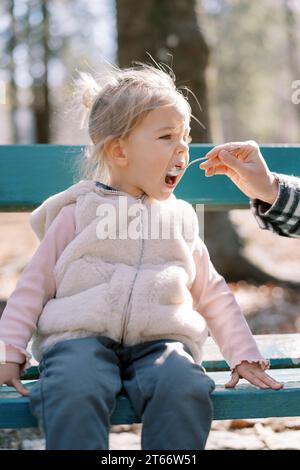 Das kleine Mädchen isst Haferbrei aus einem Löffel, den ihre Mutter sie füttert, sitzend auf einer Bank im Park. Abgeschnitten Stockfoto