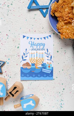 Grußkarte mit Text HAPPY HANNUKAH, Dreidels und Kartoffelpfannkuchen auf weißem Tisch Stockfoto