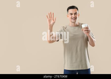 Junger Mann mit Kaffeetasse, der Hand auf beigefarbenem Hintergrund winkt Stockfoto