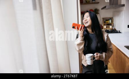 Junge schöne Frau, die einen Kaffee trinkt und eine Sprachnachricht mit einem roten Telefon neben einem Fenster in einer Küche sendet. Lächelnde Frau, die ein Kaffeebrot genießt Stockfoto