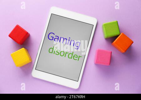 Tablette mit Phrase Gaming Disorder und bunten Würfeln auf violettem Hintergrund, flach gelegt. Suchtverhalten Stockfoto