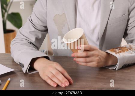 Frau zeigt Flecken von Kaffee auf ihrer Jacke an Holztisch drinnen, Nahaufnahme Stockfoto