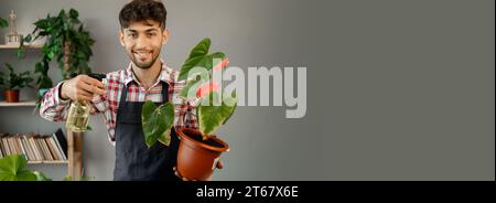 Hauspflanzenpflegekonzept. Arabischer männlicher Gärtner, der Spathiphyllum-Pflanze mit Sprühflasche bewässert, die in der Werkstatt arbeitet und in die Kamera blickt. Pflanzen von Zuhause Stockfoto