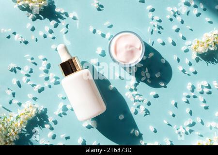 Kosmetisches Serum in weißer Flasche ohne Marke und Cremebecher. Blauer Hintergrund mit kleinen weißen Blumen und Blütenblättern. Starke Schatten. Draufsicht, flach. Skinc Stockfoto